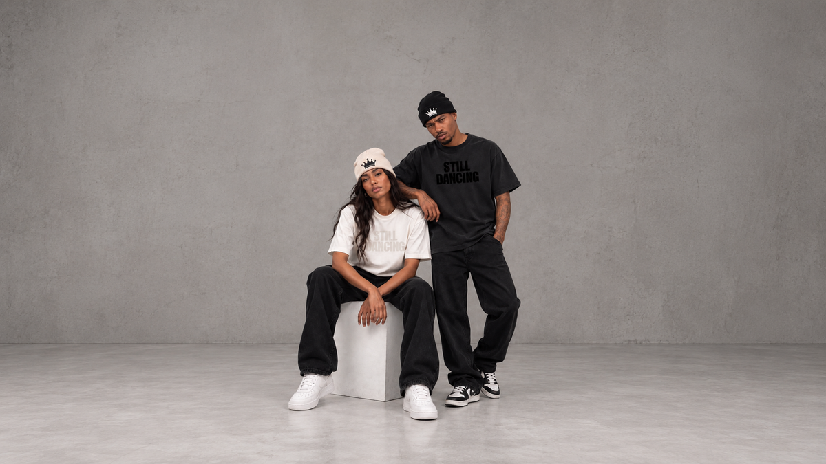Two models posing in a minimalist studio setting with a textured gray background. One model is seated on a white block, wearing a white t-shirt, black sweatpants, white sneakers, and a beige baseball cap.